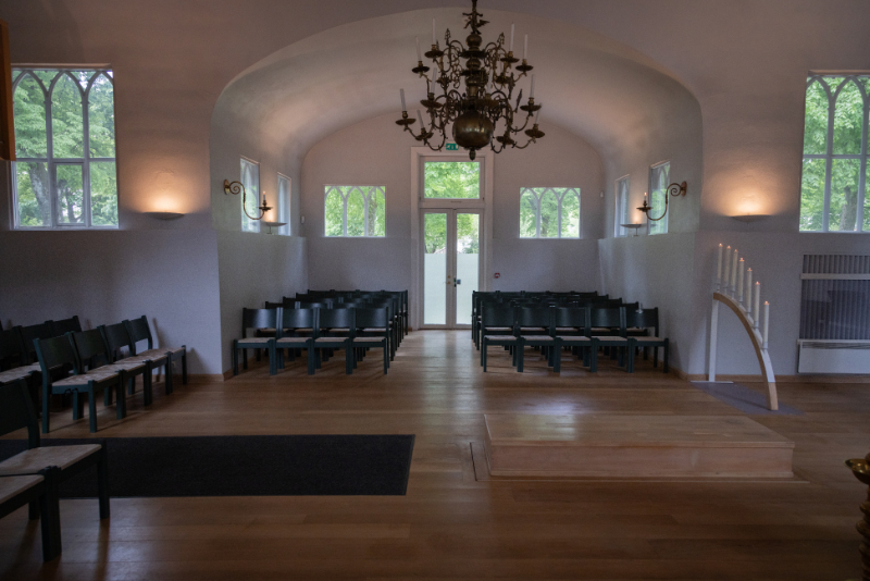 Interiørbilde fra Kristiansand kapell, som viser det enkle og elegante rommet med buede vegger, lyse overflater og grønne stoler plassert symmetrisk. En stor lysekrone henger i midten, og lys fra de gotiske vinduene skaper en fredelig atmosfære. Til høyre står en lysbue med tente lys som tilfører en høytidelig stemning. Foto.