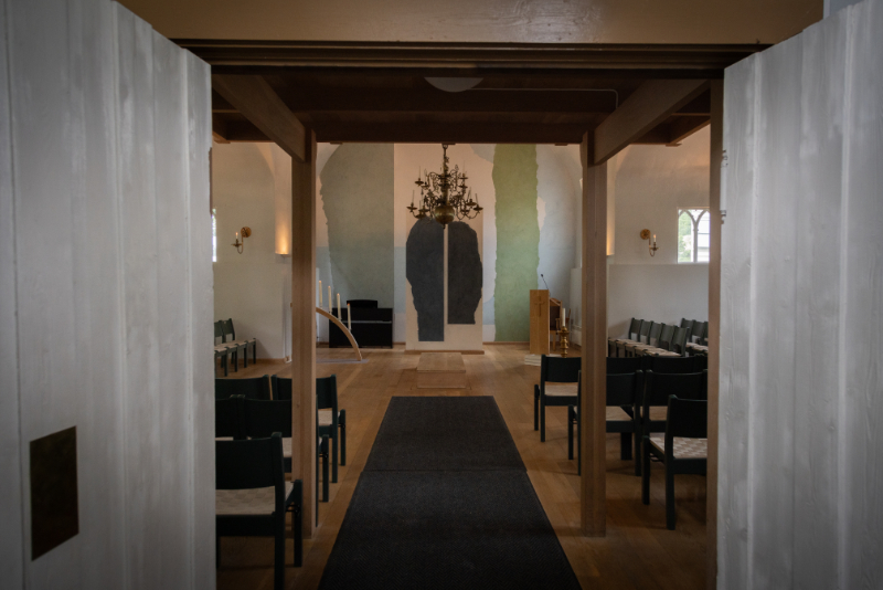 Interiørbilde fra Kristiansand kapell, sett gjennom dørene inn mot hovedrommet. Kapellet har et minimalistisk og moderne design, med en lysekrone sentralt plassert over midtgangen. Veggene er dekorert med abstrakte fargetoner i grønt og blått, som skaper et rolig og kunstnerisk uttrykk. Stolene er enkelt plassert, og til venstre står en lysbue som tilfører en høytidelig stemning. Foto.