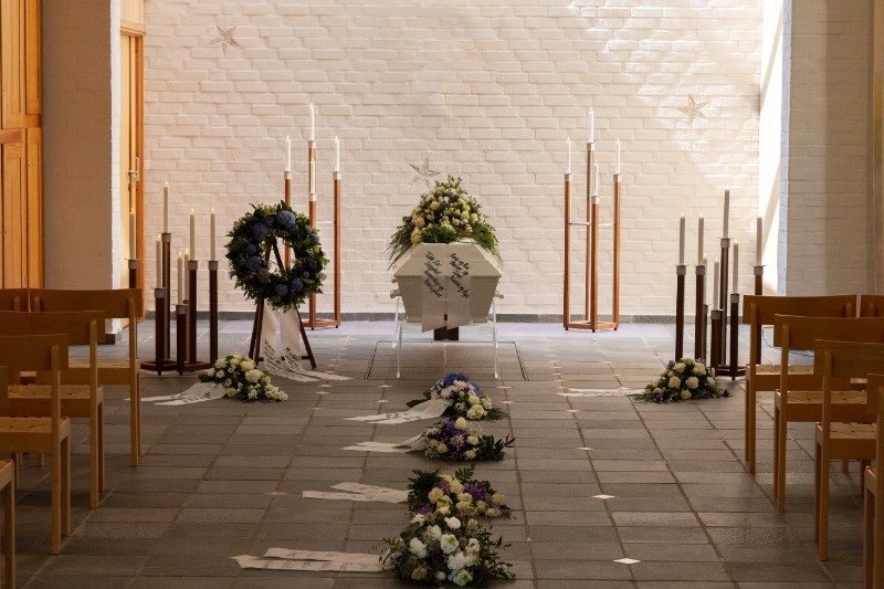 En dekorert kiste i midten av seremonilokalet i Oddernes kapell, omgitt av blomsteroppsatser, kranser og tente lys. Lyse vegger og minimalistisk interiør gir en rolig og høytidelig atmosfære. Foto.
